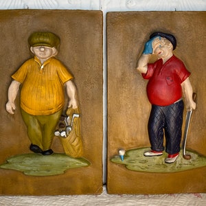 Op de afbeelding: Twee keramische wandplaquettes die golfers afbeelden. De golfer links draagt een geel shirt, een groene broek en een bruine pet. Hij draagt een golftas met clubs. De golfer rechts draagt een rood shirt, een blauwe broek en een blauwe pet. Hij houdt een golfclub vast en veegt zijn voorhoofd af met een blauwe handdoek.