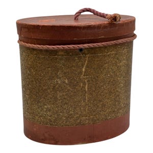 May include: An oval-shaped hat box with a brown and green marbled paper exterior. The box has a red paper lid with a rope handle.
