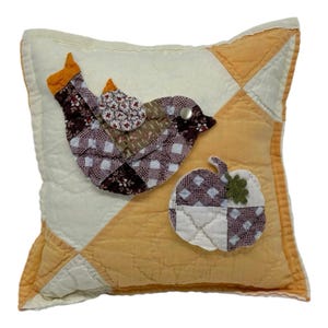 May include: A square quilted throw pillow featuring a patchwork bird and a pumpkin appliqué. The pillow has a cream and orange geometric background. The bird is crafted from brown, white, and floral fabric patches with a silver stud eye.