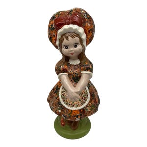 Puede incluir: Una figura de cerámica de una niña con un vestido de estampado floral marrón y naranja, un delantal blanco con volantes, un gorro a juego con un lazo rojo grande y zapatos marrones, sobre una base circular verde con textura de hierba.