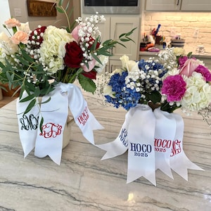 Puede incluir: Dos ramos de flores frescas con cintas blancas. Uno presenta flores rojas, blancas y rosas, mientras que el otro tiene flores azules, rosas y blancas. Las cintas están bordadas con iniciales y "HOCO 2025".