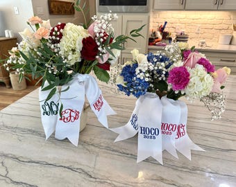 Puede incluir: Dos ramos de flores frescas con cintas blancas. Uno presenta flores rojas, blancas y rosas, mientras que el otro tiene flores azules, rosas y blancas. Las cintas están bordadas con iniciales y "HOCO 2025".