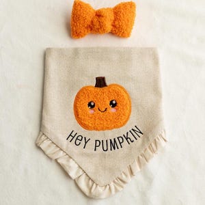 May include: A beige fabric dog bandana with a fluffy orange pumpkin and the text "Hey Pumpkin". The bandana has a ruffled edge and comes with a matching orange fluffy bow tie.