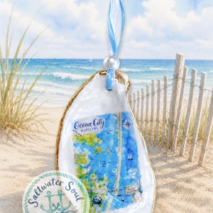 May include: A decorative oyster shell ornament featuring a watercolor map of Ocean City, Maryland. The shell has a gold-painted edge and is suspended by a light blue and white striped ribbon with a single white pearl bead accent at the top.