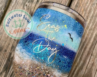 Könnte beinhalten: Ein blauer und goldener Glitzerbecher mit dem Text "Seas the Day" und einem Möwenmotiv. Der Becher hat eine weiße und braune Glitzer-Schicht am Boden, die Sand ähnelt.