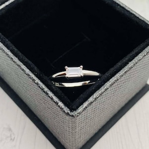 May include: A silver ring with a rectangular clear stone set in the center. The ring is in a black velvet box.