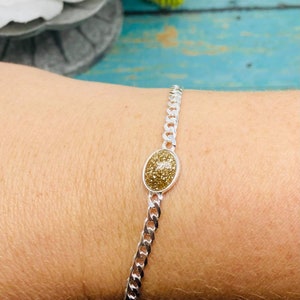 May include: A silver-toned chain bracelet featuring a central oval-shaped charm with a textured, shimmering gold-colored surface. The bracelet consists of interlocking metal links that form a continuous band around the wrist, showcasing a metallic finish.