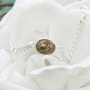 May include: A silver-toned chain bracelet featuring a central oval-shaped charm with a textured, shimmering gold-colored surface. The bracelet is composed of small, interlocking metal links that connect to the central oval piece on both sides.