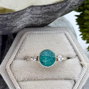 May include: A silver ring with a round, teal-colored stone in a bezel setting. Two smaller, clear stones flank the main stone. The ring is displayed in a gray velvet ring box.