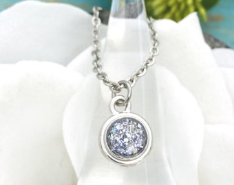 May include: A silver-toned metal necklace featuring a circular pendant with a textured, light blue, glittery center. The pendant hangs from a delicate link chain, showcasing a reflective, sparkling surface within a smooth, polished metal bezel setting.
