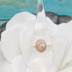 May include: A gold ring with a white oval-shaped stone embedded with gold flecks. The ring is displayed on a white flower.