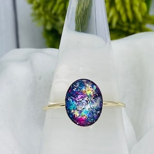 May include: A gold ring with a colorful oval-shaped resin stone. The stone has a blue, purple, and pink color scheme with iridescent glitter.