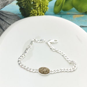 May include: A silver chain bracelet with a small oval-shaped charm. The charm is covered in tiny gold glitter.