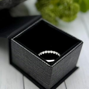 May include: A diamond eternity ring rests inside a black textured jewelry box with a black velvet interior. The ring is set with multiple small, round diamonds. The box is open, revealing the ring.