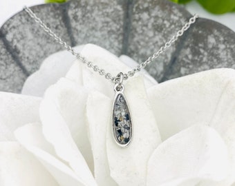 May include: A silver-toned necklace featuring a teardrop-shaped pendant. The pendant has a polished metal border and is filled with a textured inlay of small, multi-colored flecks in shades of black, white, and gray, suspended on a delicate chain.