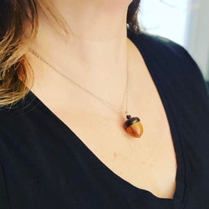 May include: A silver chain necklace with a wooden acorn pendant. The acorn is brown with a black top.