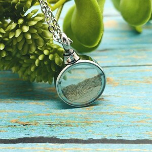 May include: A silver chain necklace with a round, clear glass pendant containing sand. The pendant is attached to a silver chain.