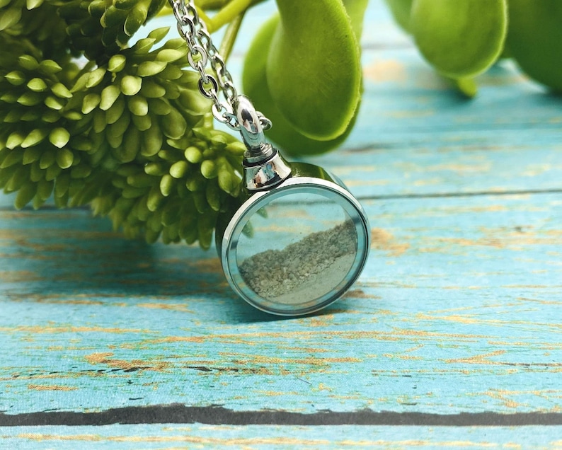 May include: A silver chain necklace with a round, clear glass pendant containing sand. The pendant is attached to a silver chain.