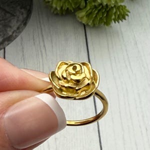 以下が含まれることがあります： 金色のリングで、バラのデザインが施されています。バラは詳細な花びらのデザインで、リングのバンドはシンプルなゴールドです。リングは人の手で持たれています。ジュエリーです。