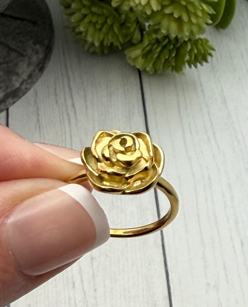 以下が含まれることがあります： 金色のリングで、バラのデザインが施されています。バラは詳細な花びらのデザインで、リングのバンドはシンプルなゴールドです。リングは人の手で持たれています。ジュエリーです。
