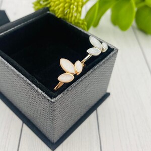 K&ouml;nnte beinhalten: Zwei goldene Ringe mit wei&szlig;en Emaille-Bl&auml;tterdesigns in einer grauen Samt-Ringbox.