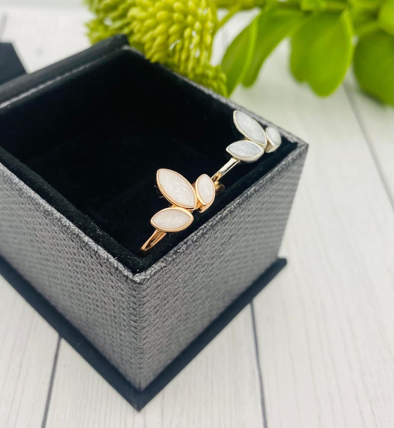 K&ouml;nnte beinhalten: Zwei goldene Ringe mit wei&szlig;en Emaille-Bl&auml;tterdesigns in einer grauen Samt-Ringbox.