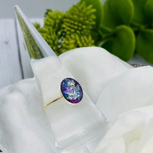 May include: A gold ring with a purple, blue, and white oval-shaped resin stone. The stone is filled with iridescent glitter.
