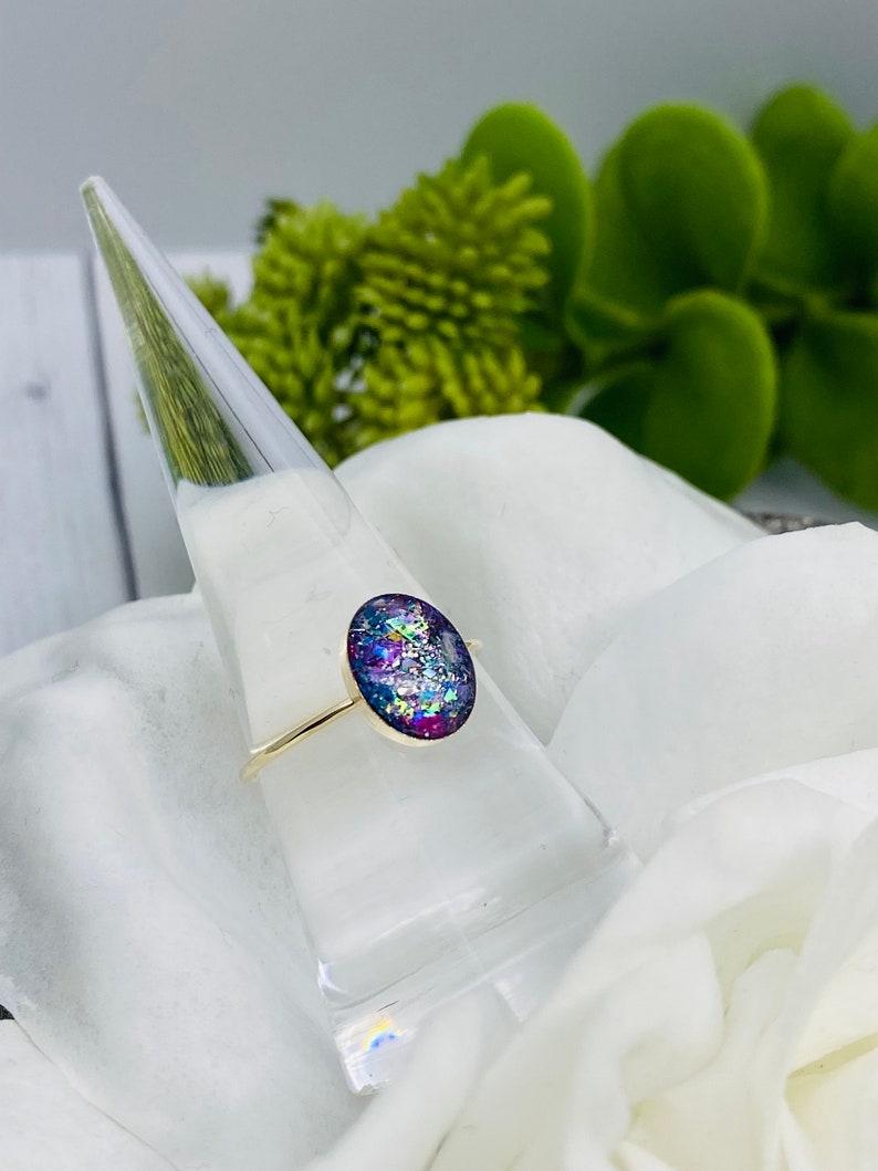 May include: A gold ring with a purple, blue, and white oval-shaped resin stone. The stone is filled with iridescent glitter.
