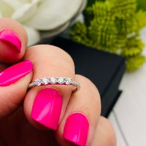 May include: A silver band ring with five small round clear stones set in a row. The ring is on a finger with bright pink nail polish.