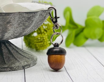 May include: A wooden acorn keychain with a silver metal loop and clasp. The acorn is brown with a dark brown cap.