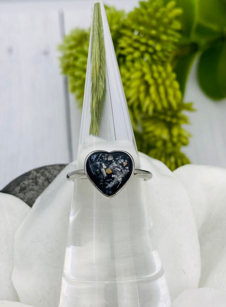 May include: A silver heart-shaped ring with a black resin inlay containing small, colorful particles. The ring is displayed on a clear ring holder.
