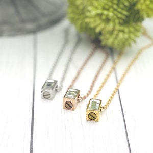 May include: Three necklaces with square pendants featuring a clear crystal and a small screw detail. The necklaces are silver, rose gold, and gold.