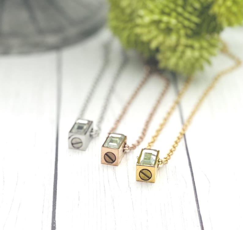 May include: Three necklaces with square pendants featuring a clear crystal and a small screw detail. The necklaces are silver, rose gold, and gold.