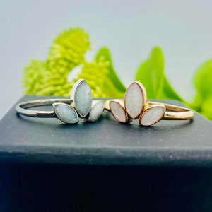 K&ouml;nnte beinhalten: Zwei Ringe aus Silber und Gold mit wei&szlig;en Steinbes&auml;tzen in Form von Bl&auml;ttern. Die Ringe werden auf einer schwarzen Schachtel mit gr&uuml;nem Blattwerk im Hintergrund pr&auml;sentiert.