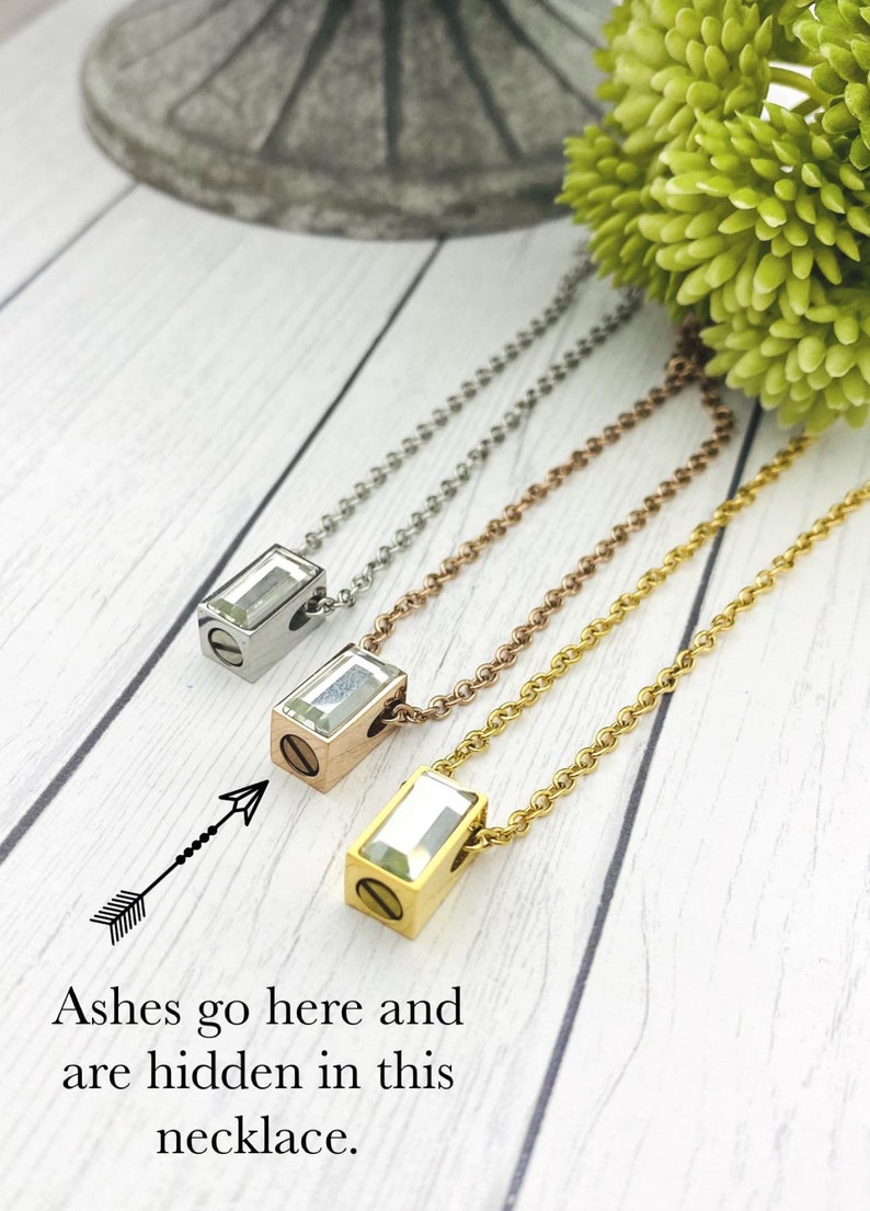May include: Three necklaces with different colored chains and a clear crystal pendant. The pendant has a screw on the side, and the text "Ashes go here and are hidden in this necklace." is below the image.