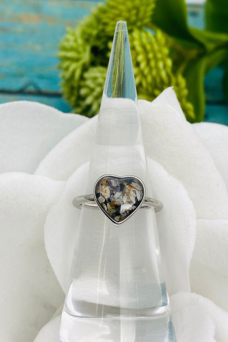 May include: A silver heart-shaped ring with a clear resin insert containing small white and brown pieces. The ring is on a clear cone-shaped ring holder.