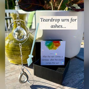 May include: A green ceramic urn with a teardrop-shaped pendant and a crystal. A black box contains a card with a rainbow design and the text: "After the rain comes a rainbow... after the storm comes the sun."