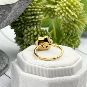 以下が含まれることがあります： 白いベルベットのリングボックスにセットされた、花の模様のゴールドリング。リングのバンドは細くて円形で、花の要素は明るい色です。背景には緑の葉が見えます。