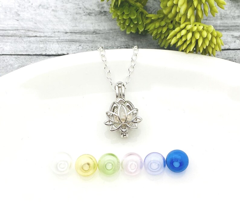 K&ouml;nnte beinhalten: Eine silberne Lotusbl&uuml;ten-Anh&auml;ngerkette mit einer klaren Glaskugel im Inneren. Die Kette wird auf einer wei&szlig;en Oberfl&auml;che mit verschiedenen farbigen Glasperlen pr&auml;sentiert.