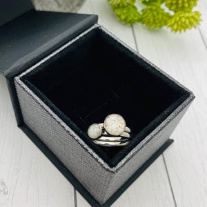 May include: A set of two silver rings with round, light-colored stone settings, presented in an open black jewelry box with a textured, gray exterior. The rings are stacked, and the box is on a light-colored surface.