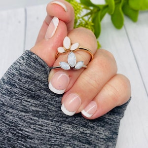 K&ouml;nnte beinhalten: Zwei Ringe mit wei&szlig;en Blumendesigns. Die Ringe befinden sich an einem Finger mit wei&szlig;er French Manik&uuml;re.