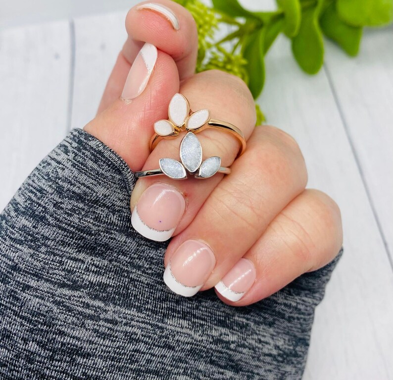 K&ouml;nnte beinhalten: Zwei Ringe mit wei&szlig;en Blumendesigns. Die Ringe befinden sich an einem Finger mit wei&szlig;er French Manik&uuml;re.