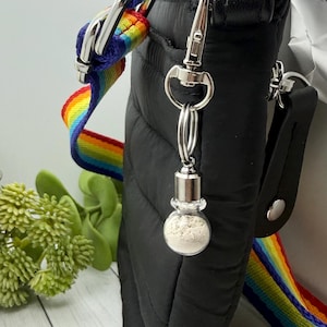 May include: A black quilted crossbody bag featuring a vibrant rainbow-striped adjustable strap and a silver-toned metal lobster clasp. Attached to the clasp is a small, clear glass spherical charm containing a fine, light-colored powdery substance.