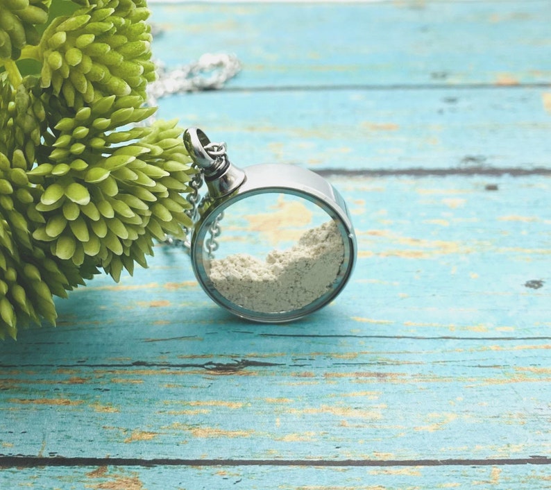 May include: A silver necklace with a round pendant containing a white powder. The pendant is open on one side, revealing the powder inside.