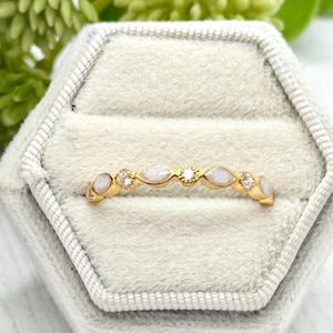 May include: A gold ring with a delicate design. The ring features alternating oval-shaped white stones and small, round clear stones. The ring is set in a gold band and is displayed in a gray ring box.