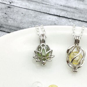 K&ouml;nnte beinhalten: Zwei silberne Ketten mit unterschiedlichen Anh&auml;ngerdesigns. Ein Anh&auml;nger ist eine Lotusblume mit einer gr&uuml;nen Glasperle im Inneren. Der andere Anh&auml;nger ist ein spiralf&ouml;rmiger K&auml;fig mit einer gelben Glasperle im Inneren. Im Hintergrund befinden sich vier Glasperlen, eine klare, eine gelbe, eine gr&uuml;ne und eine rosa.
