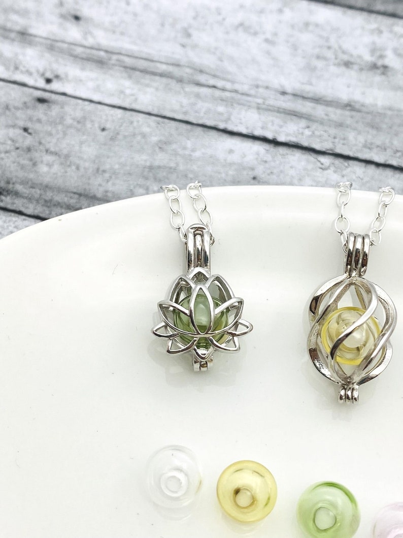 K&ouml;nnte beinhalten: Zwei silberne Ketten mit unterschiedlichen Anh&auml;ngerdesigns. Ein Anh&auml;nger ist eine Lotusblume mit einer gr&uuml;nen Glasperle im Inneren. Der andere Anh&auml;nger ist ein spiralf&ouml;rmiger K&auml;fig mit einer gelben Glasperle im Inneren. Im Hintergrund befinden sich vier Glasperlen, eine klare, eine gelbe, eine gr&uuml;ne und eine rosa.