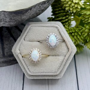 May include: Two oval-shaped opal rings with halo settings are displayed in a gray velvet ring box. One ring has a gold band, the other a silver band. The opals have a milky white color with flashes of blue and pink.