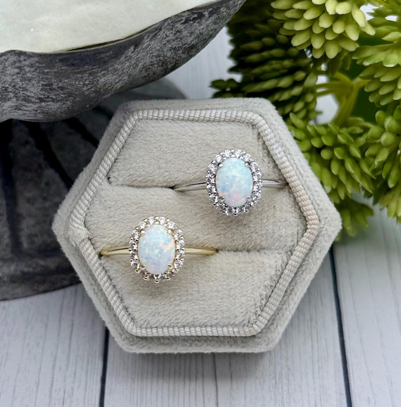 May include: Two oval-shaped opal rings with halo settings are displayed in a gray velvet ring box. One ring has a gold band, the other a silver band. The opals have a milky white color with flashes of blue and pink.