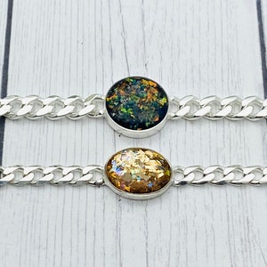 May include: Two silver-toned chain bracelets featuring central oval settings. The top bracelet contains a dark, iridescent, multi-colored flecked inlay, while the bottom bracelet features a gold-toned, shimmering, metallic-flecked inlay.